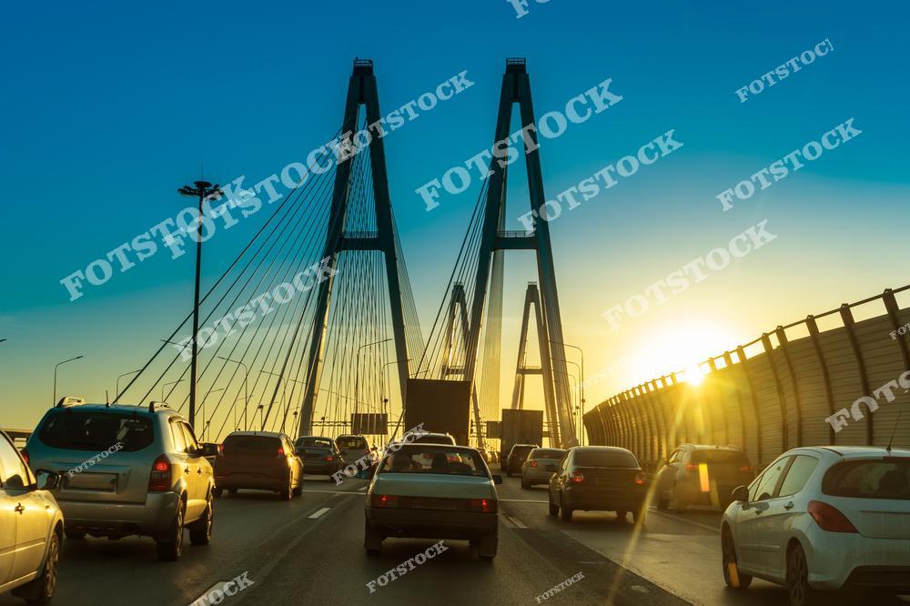 Современный вантовый мост в Санкт-Петербурге с плотным автомобильным движением в лучах вечернего солнца. Высокие пилоны, стальные тросы, городская магистраль и поток машин создают динамичный урбанистический пейзаж северной столицы России. Фотография подчёркивает красоту инженерной архитектуры, развитие транспортной инфраструктуры и атмосферу большого современного города. Изображение идеально подходит для публикаций о дорогах Санкт-Петербурга, автомобильных мостах, городском трафике, урбанистике, путешествиях и современной архитектуре России.